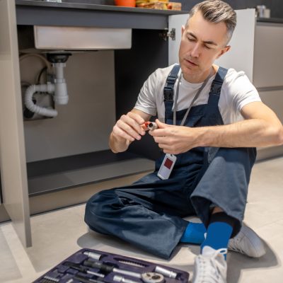 Reflections. Middle-aged man in white tshirt and blue overalls with badge sitting on floor looking at plumbing piece in hands near open set of tools in kitchen